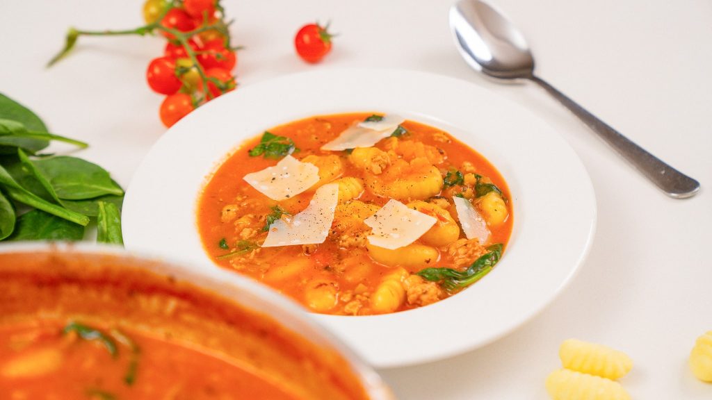 Chicken sausage gnocchi soup served in a white bowl, with spinach and cherry tomatoes artfully arranged around the dish for a fresh, vibrant look.