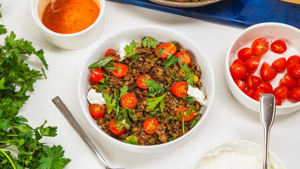 White bowl on counter with a ground beef pasta recipe- a delicious take on Turkish food. There is a bowl of cherry tomatoes on the side as well as a butter dipping sauce in a large ramekin.