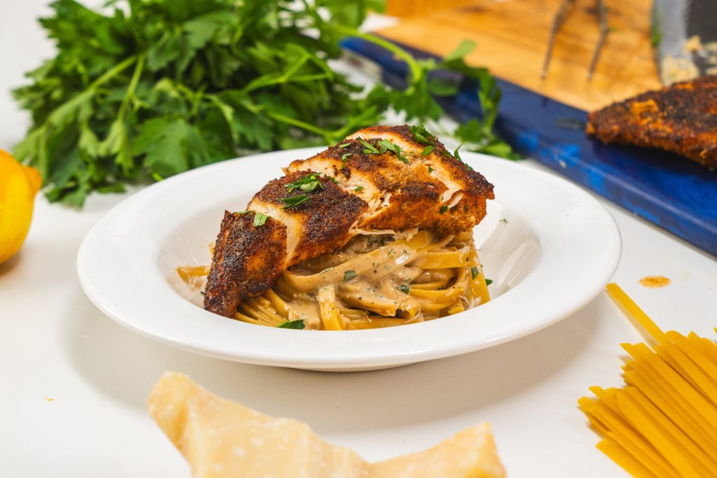 A bowl of chicken fettuccine alfredo with grilled chicken on top. Fresh parsley and a block of parmesan cheese is visible.