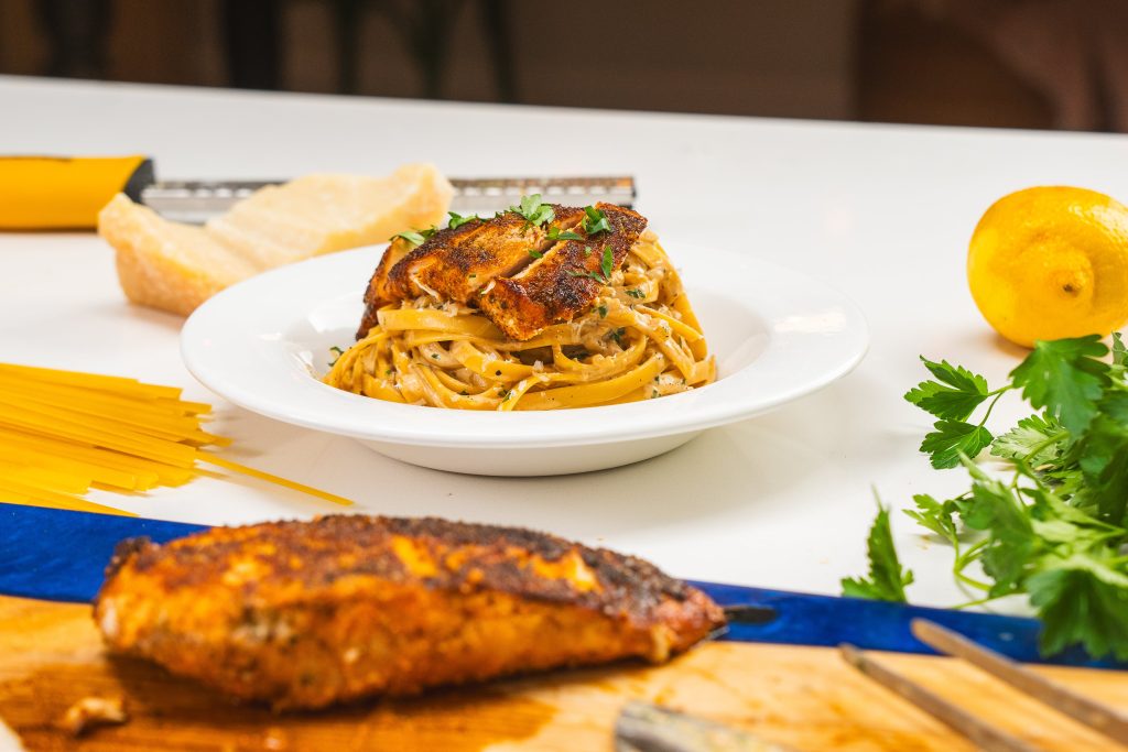 A bowl of chicken fettuccine alfredo with grilled chicken on top. Cooked chicken breast and a block of parmesan cheese is visible.