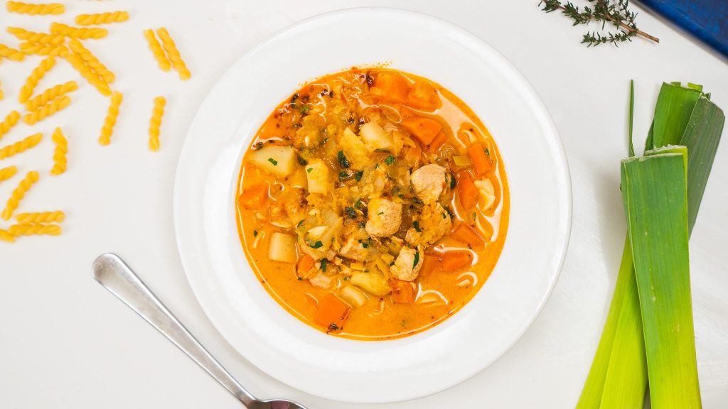 White bowl filled with spicy, creamy chicken noodle soup. There is a spoon nearby, fresh celery to the right and dried pasta to the left.