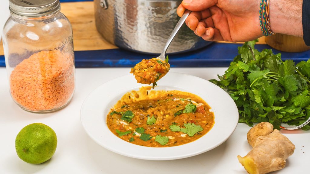 A hand holding a spoon full of red lentil dahl over a full bowl. 