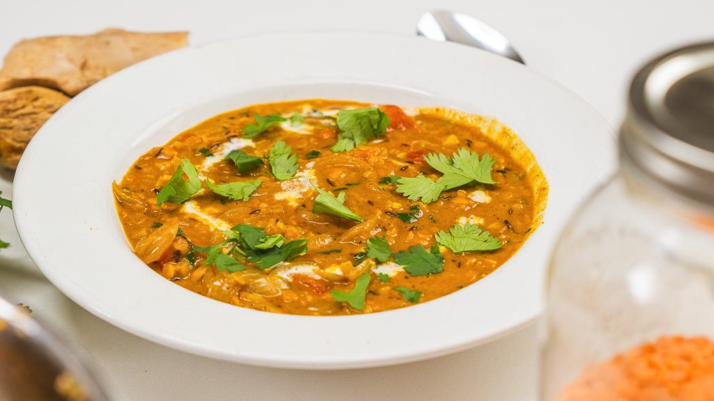 A bowl of red lentil dahl topped with fresh cilantro on a white counter with a spoon visible to the right.