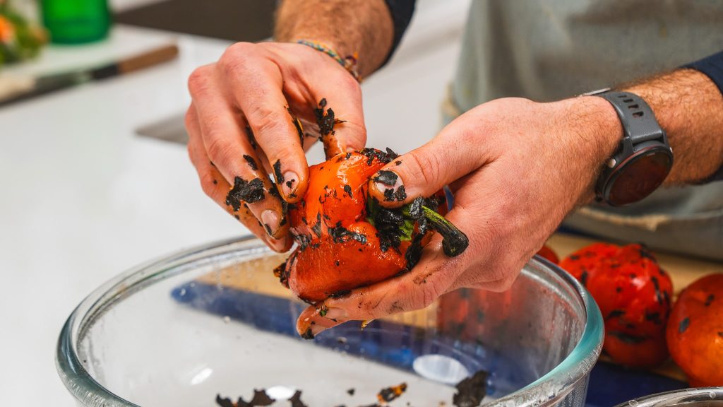 Hands peeling charred red peppers into a clear white bowl.