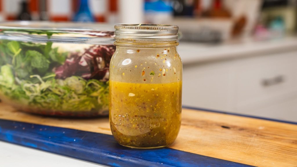 A mason jar of homemade salad dressing and a bowl of salad in the distance.