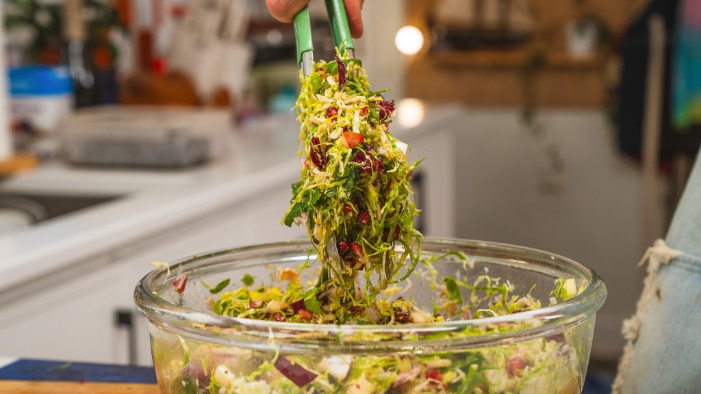 A clear bowl with colour fun salad and tongs over top scooping out some of the salad.