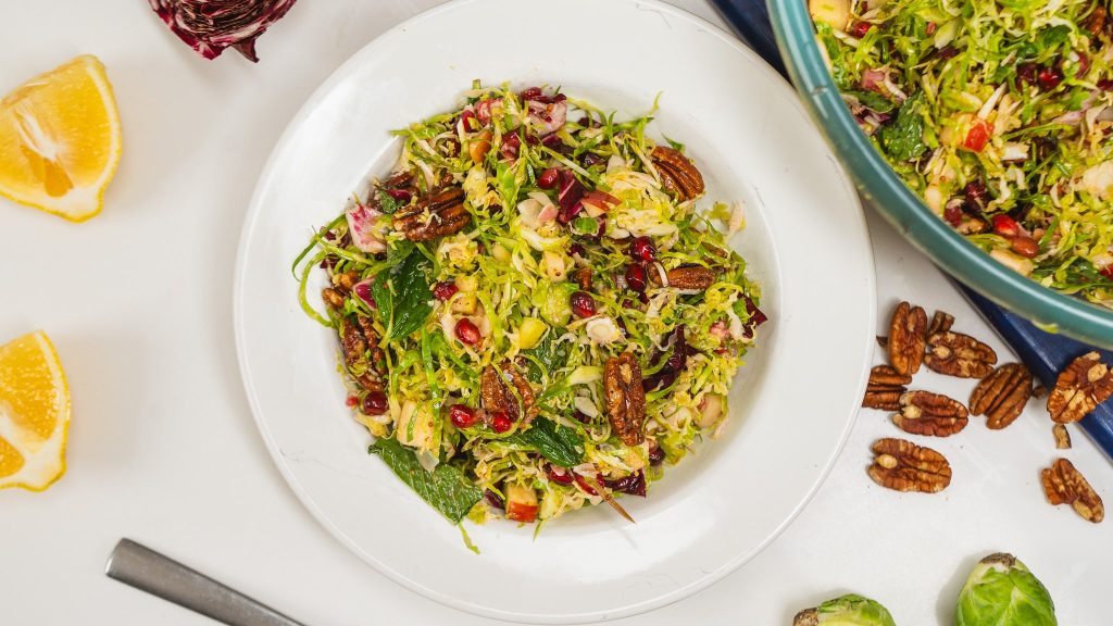 Large, white-rimmed bowl on counter with a colourful salad of brussel sprouts and other vegetables inside.