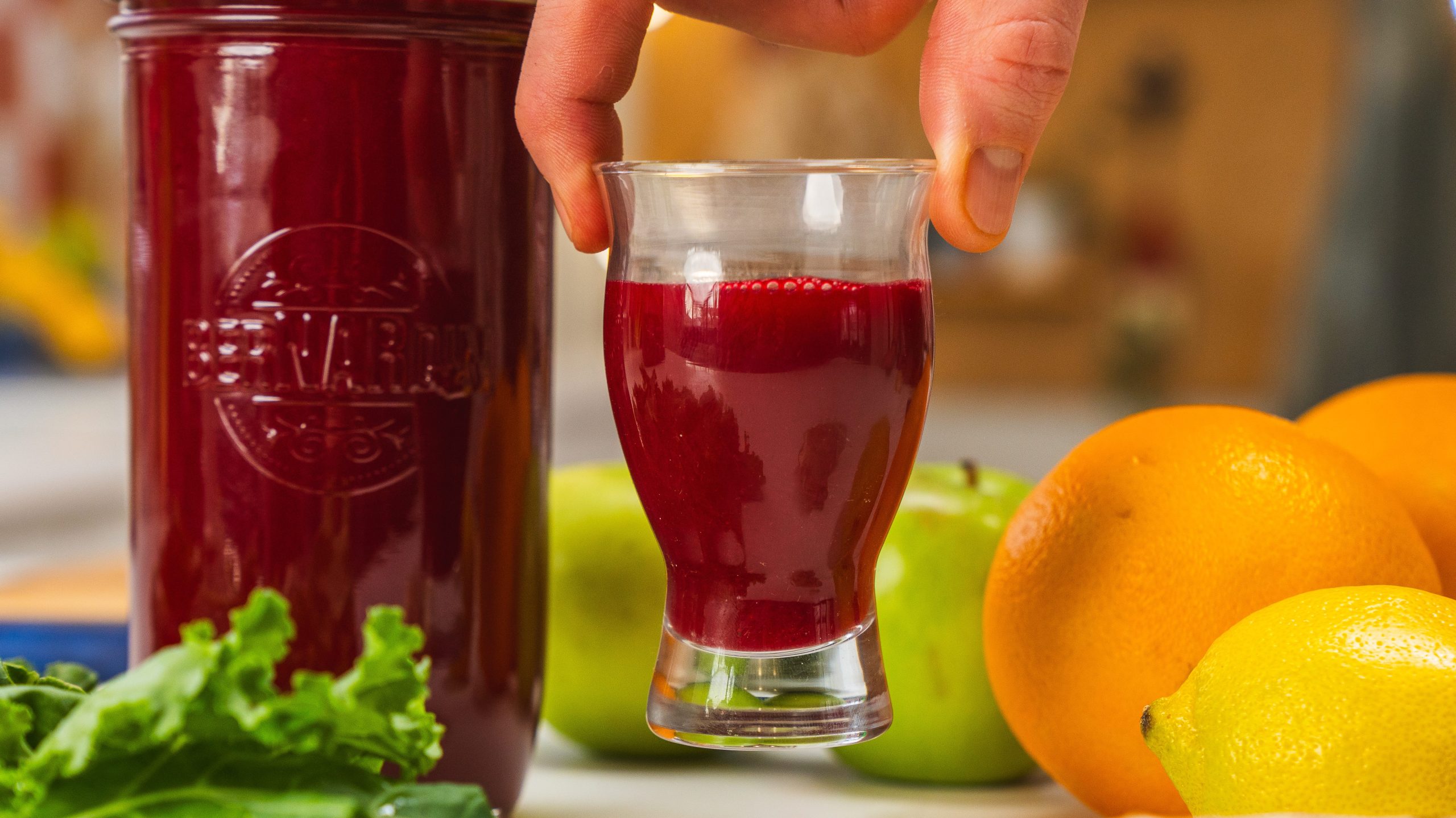 A hand holding a shot glass of beet and ginger juice. Fresh oranges, lemon and kale is visible to the side.