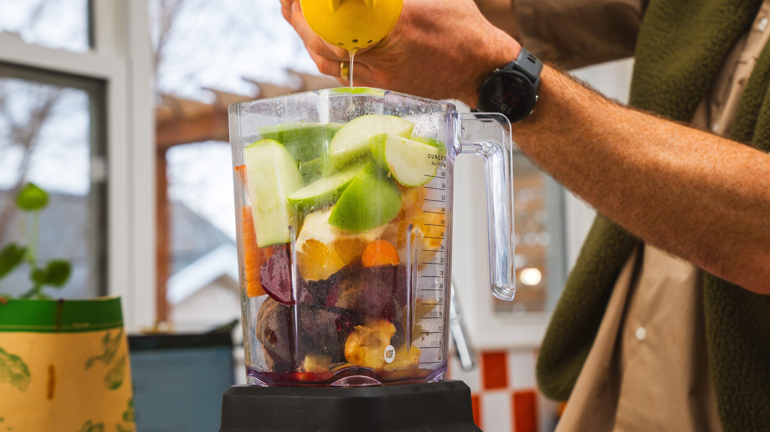 A blender filled with citrus fruit, kale, ginger and beets. A hand is squeezing fresh lemon juice in the blender. 