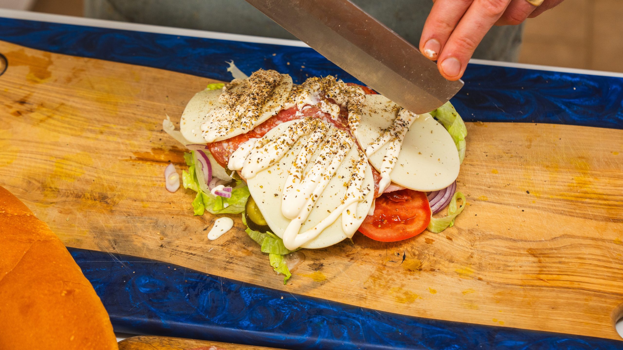 Deli meat, tomatoes, lettuce, pickles, provolone cheese and drizzled mayonnaise on a cutting board with a hand holding a knife over top. These are the ingredients to make an Italian chopped sandwich.