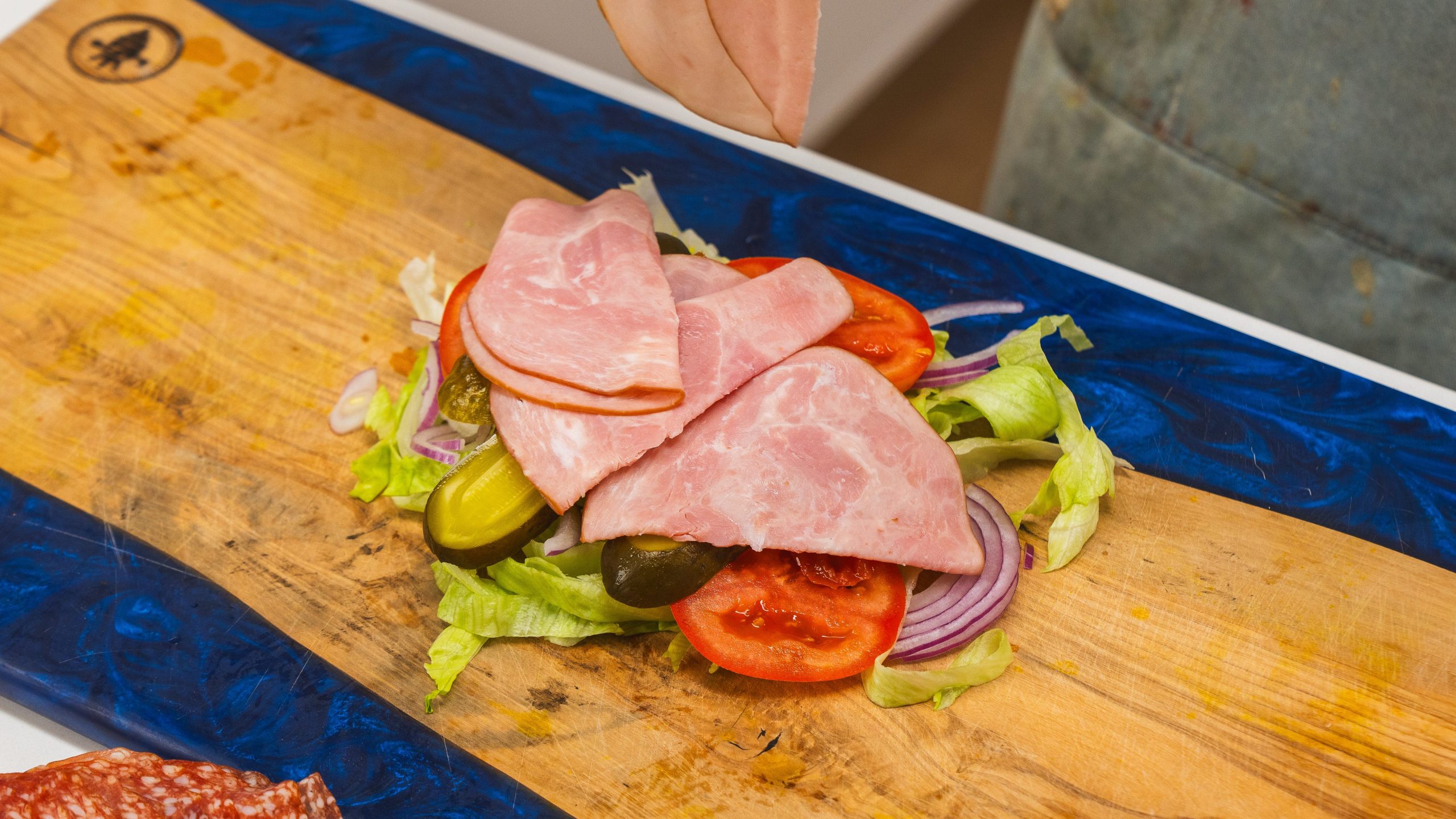 Deli meat, tomatoes, lettuce, pickles on a cutting board with a hand holding a knife over top. These are the ingredients to make an Italian chopped sandwich.
