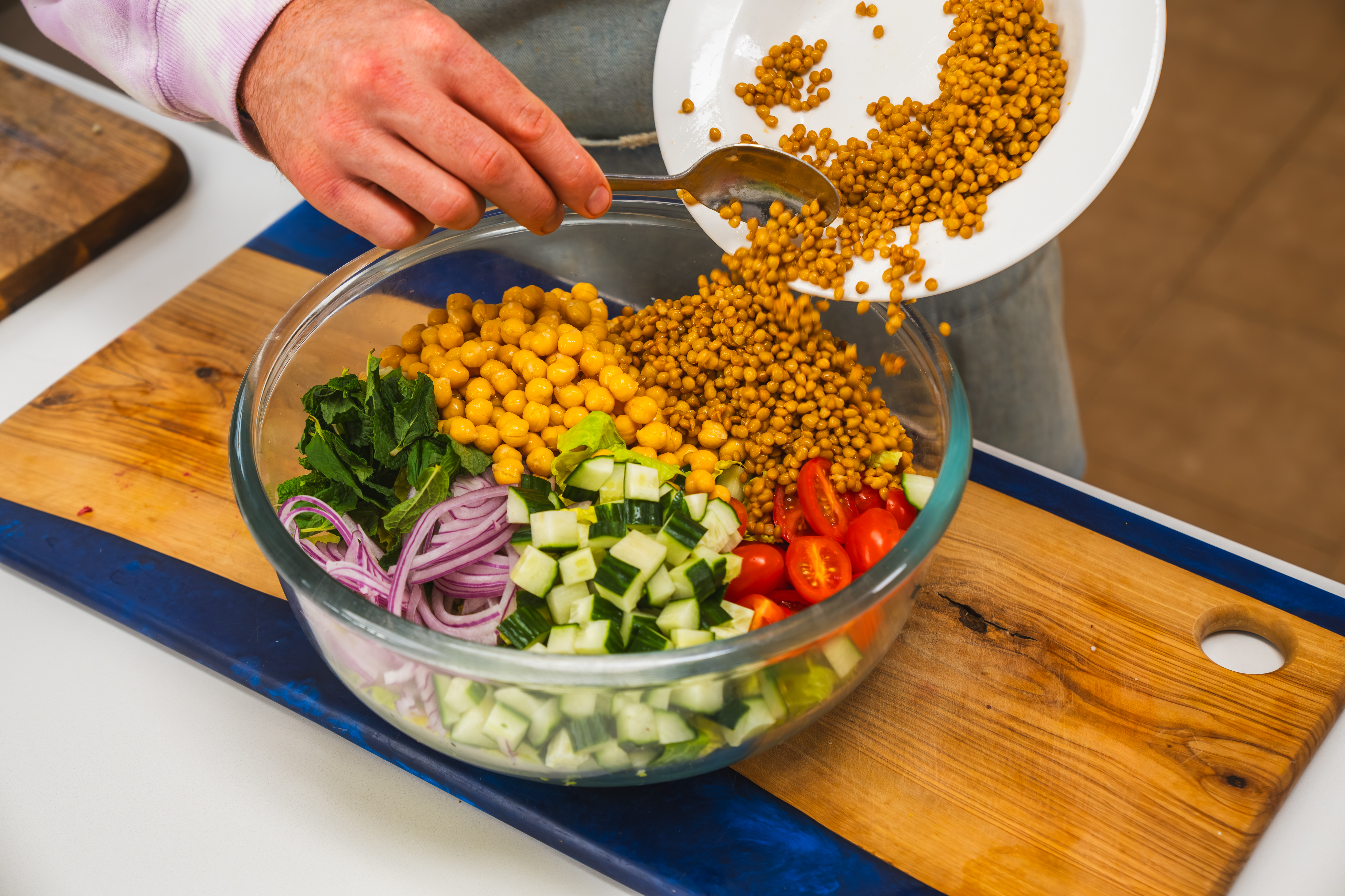 A large, clear bowl filled with a Middle Eastern Salad of fresh vegetables, chickpeas, lentils and herbs. A hand is stopping out additional lentils to add to the bowl.