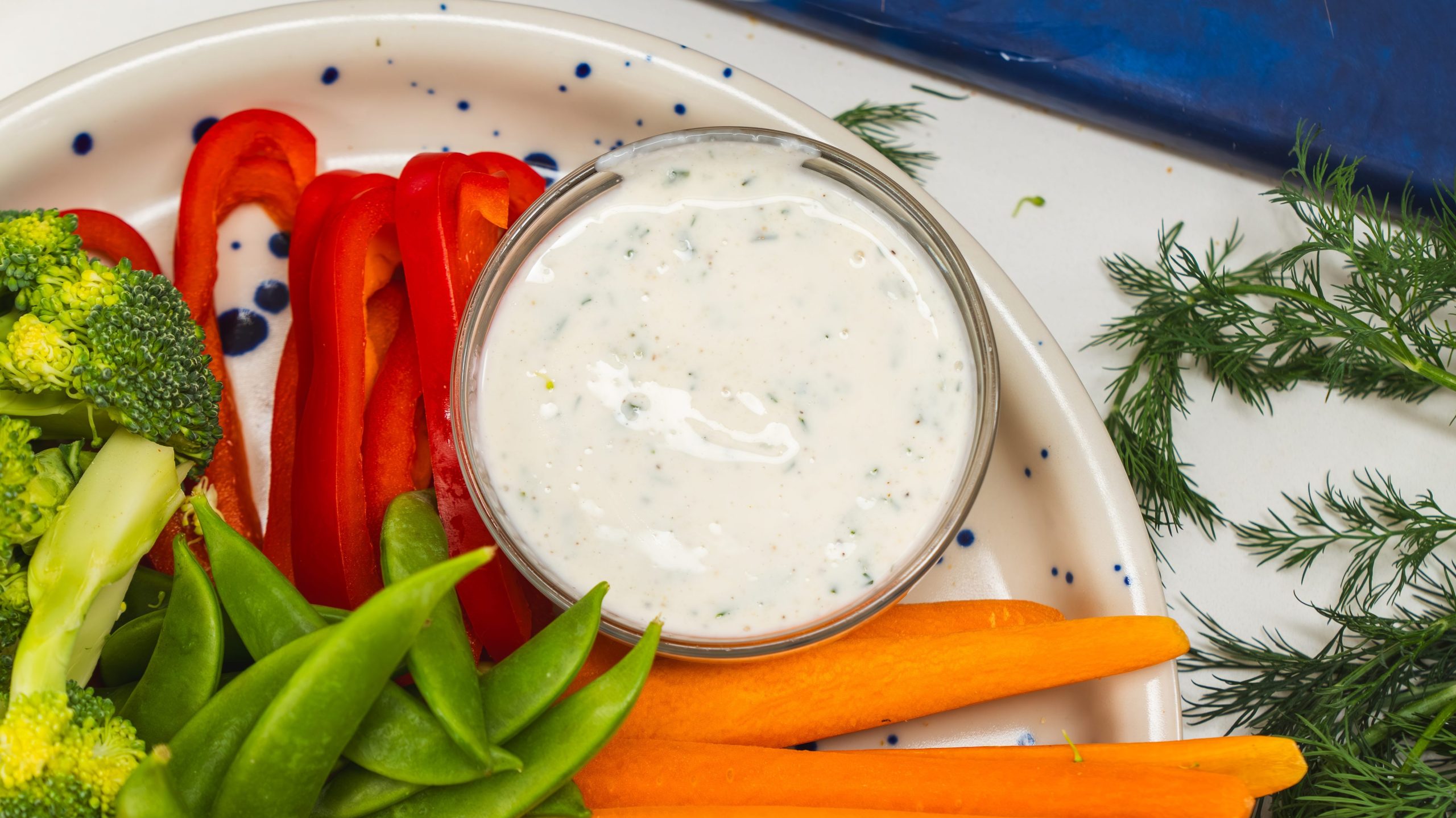 A white plate of colourful, fresh veggies and a small bowl of homemade healthy ranch dressing on the side.