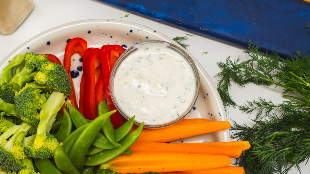 A white plate of colourful, fresh veggies and a small bowl of homemade healthy ranch dressing on the side.