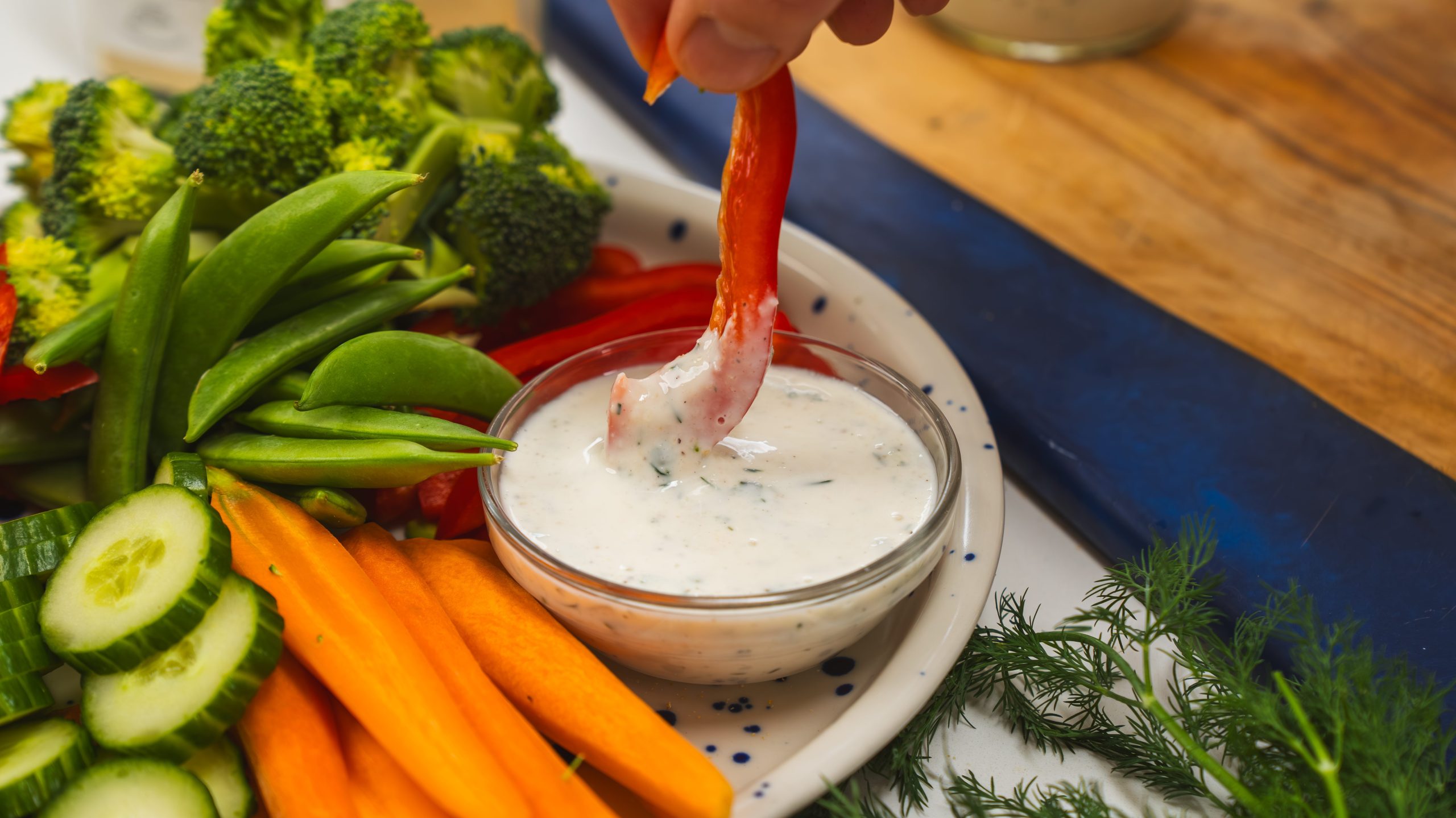 A bowl of homemade healthy ranch dressing. A hand is holding a red pepper slice and dipping it into the dressing.
