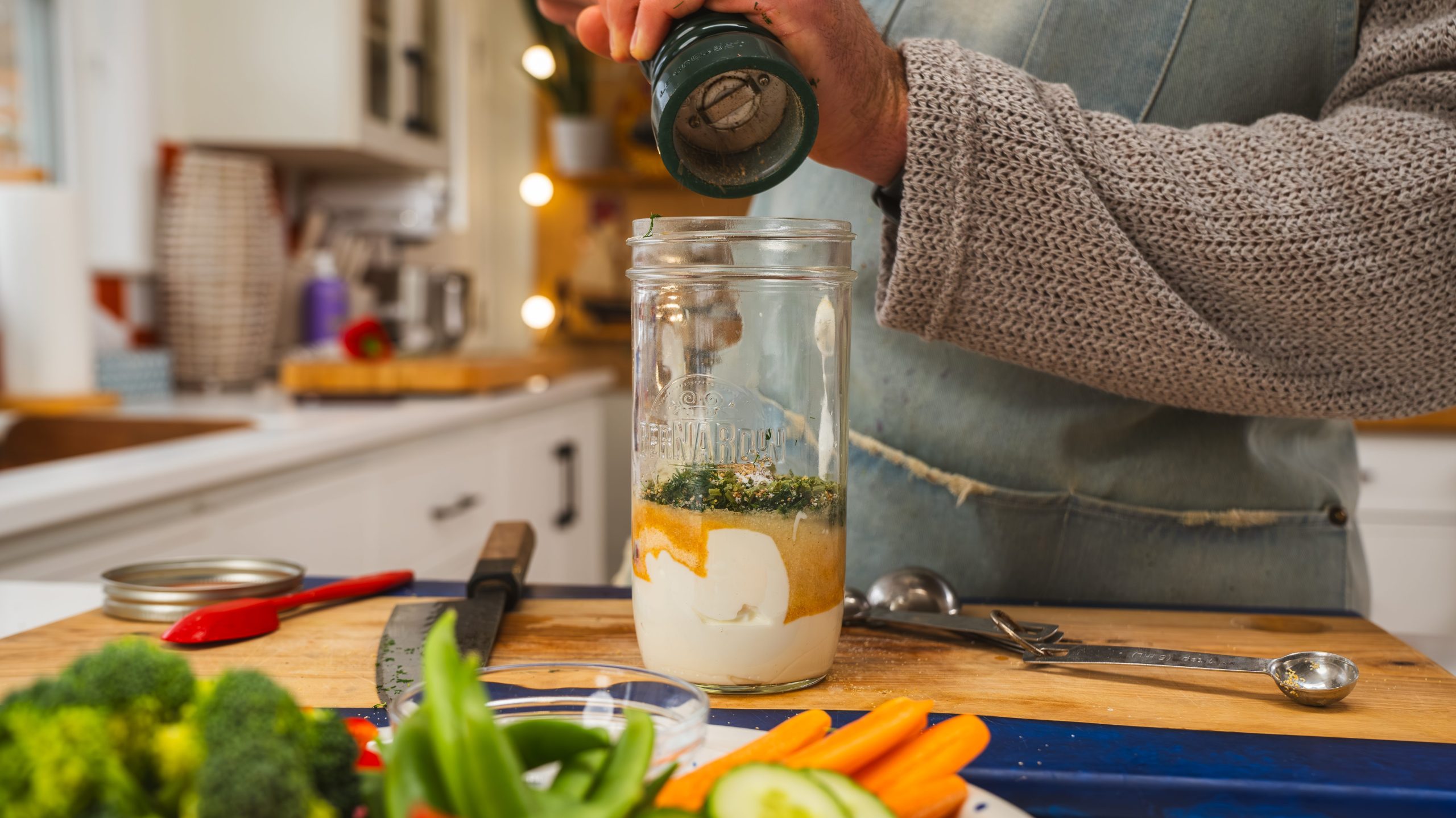 A mason jar full of homemade ranch dressing ingredients and someone grinding fresh ground pepper into the jar. Fresh vegetables are visible on a plate in front of the mason jar.