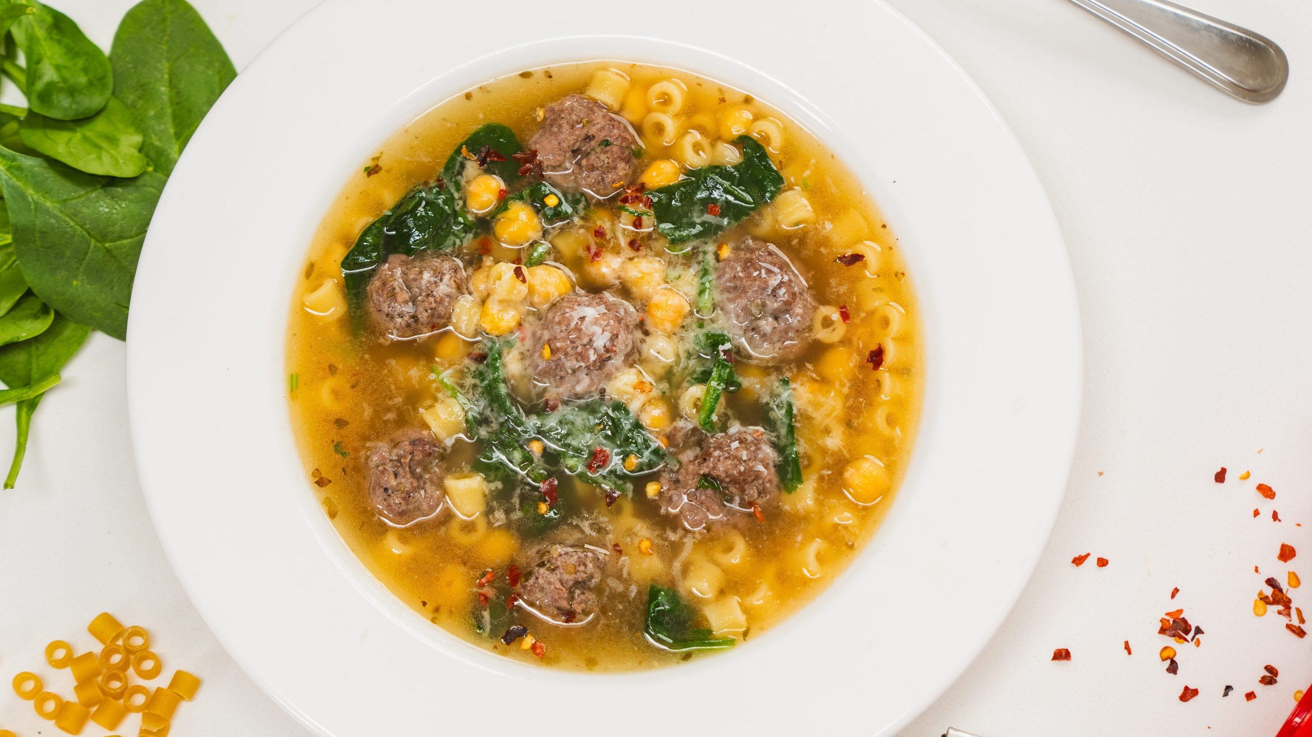 A while bowl filled with Italian Wedding Soup sitting on a white counter. A spoon as well as ry pasta and fresh spinach is visible to the side. This Italian wedding soup recipe is warm and delicious!