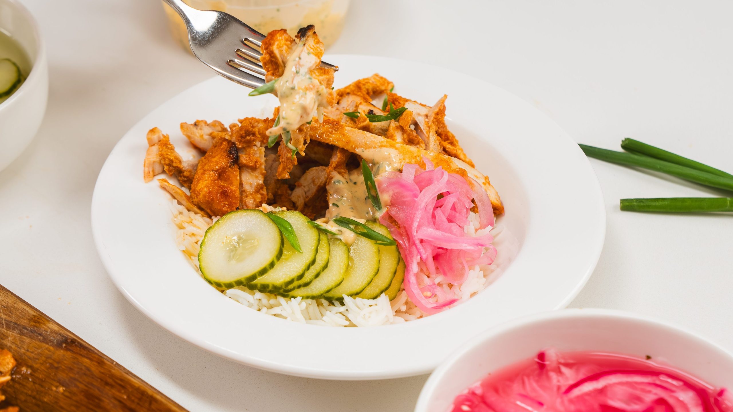 White bowl filled with loaf pan chicken shawarma, rice, slices of cucumber and pickled red onion. Visible to the side is a fork and a bowl of pickled red onion. A hand is holding a fork with some chicken.