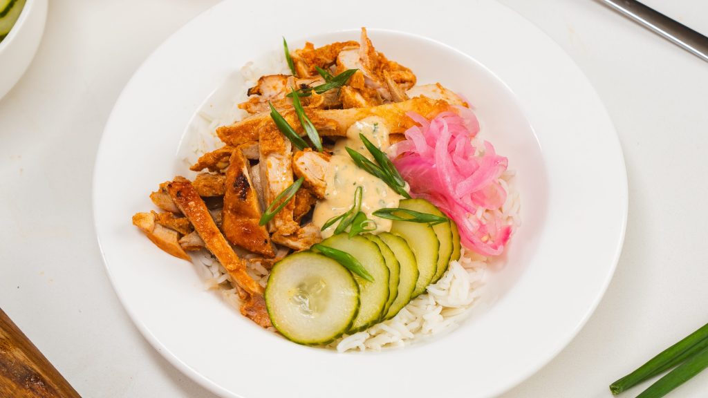 White bowl filled with loaf pan chicken shawarma, rice, slices of cucumber and pickled red onion.