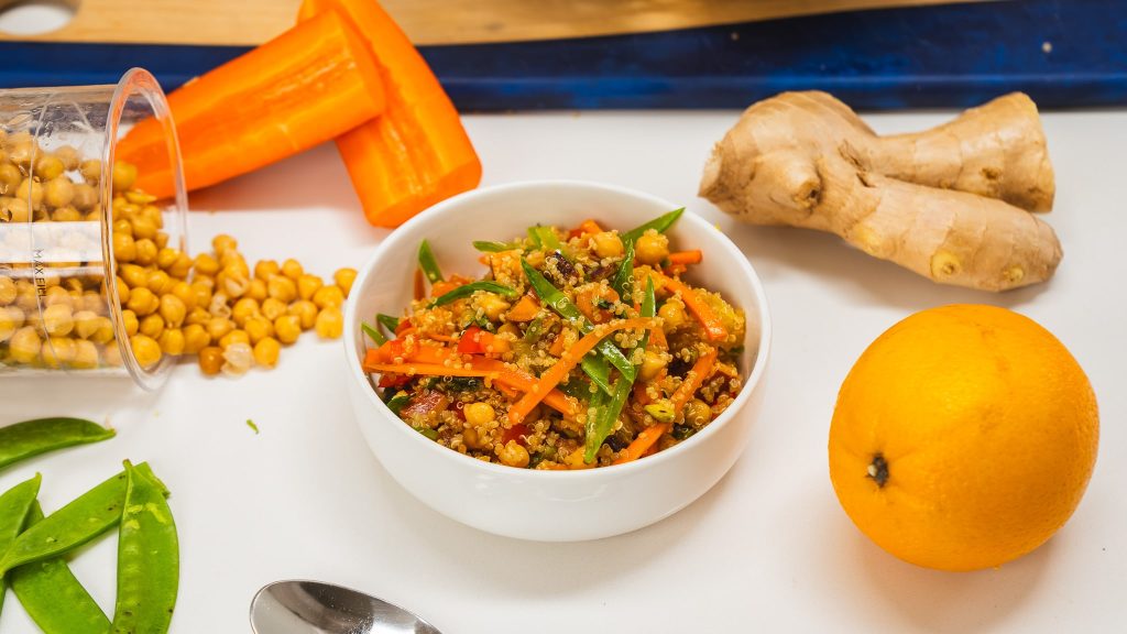 Quinoa salad in a white bowl filled with colourful vegetables. Ingredients are visible to the side-fresh ginger, peppers, and chickpeas.