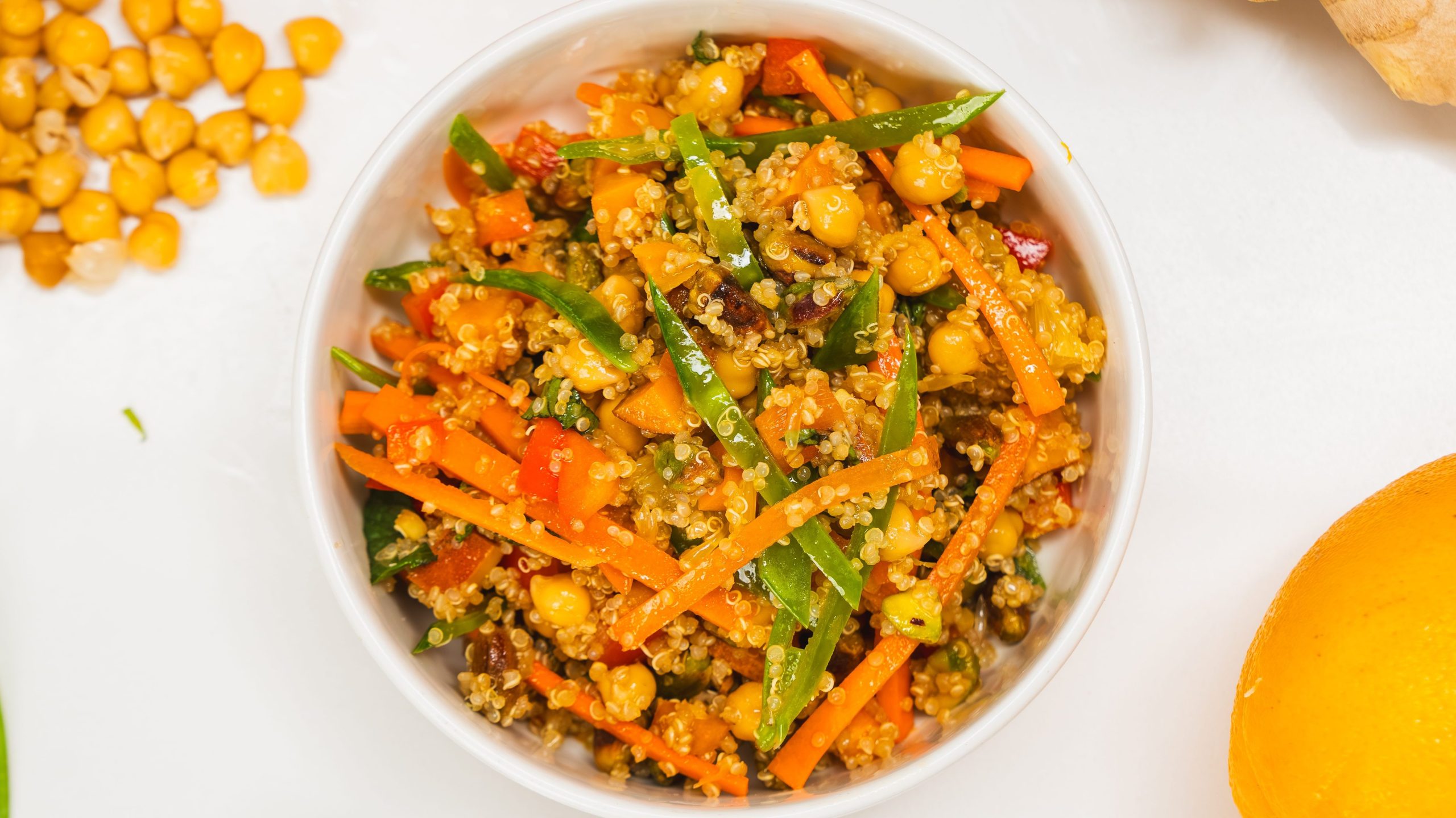 A white bowl filled with a quinoa salad and fresh, crunchy vegetables. A spoon, chickpeas, orange and ginger can be seen on the sides. This quinoa salad recipe is colourful and delicious!