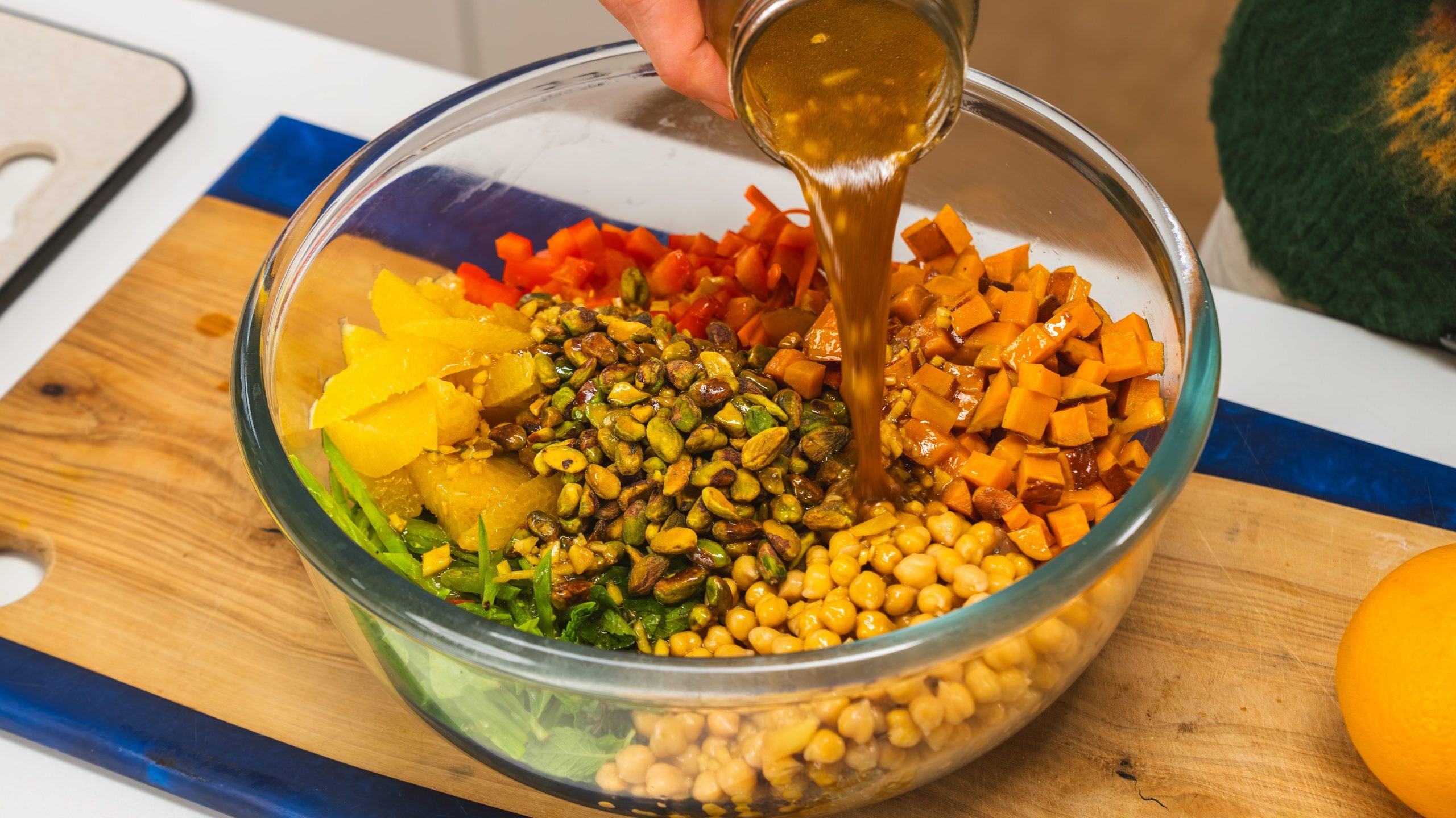 A glass bowl filled with quinoa, chickpeas, fresh vegetables carrots and red pepper. A hand above the bowl is holding a mason jar of salad dressing and is pouring it on top.