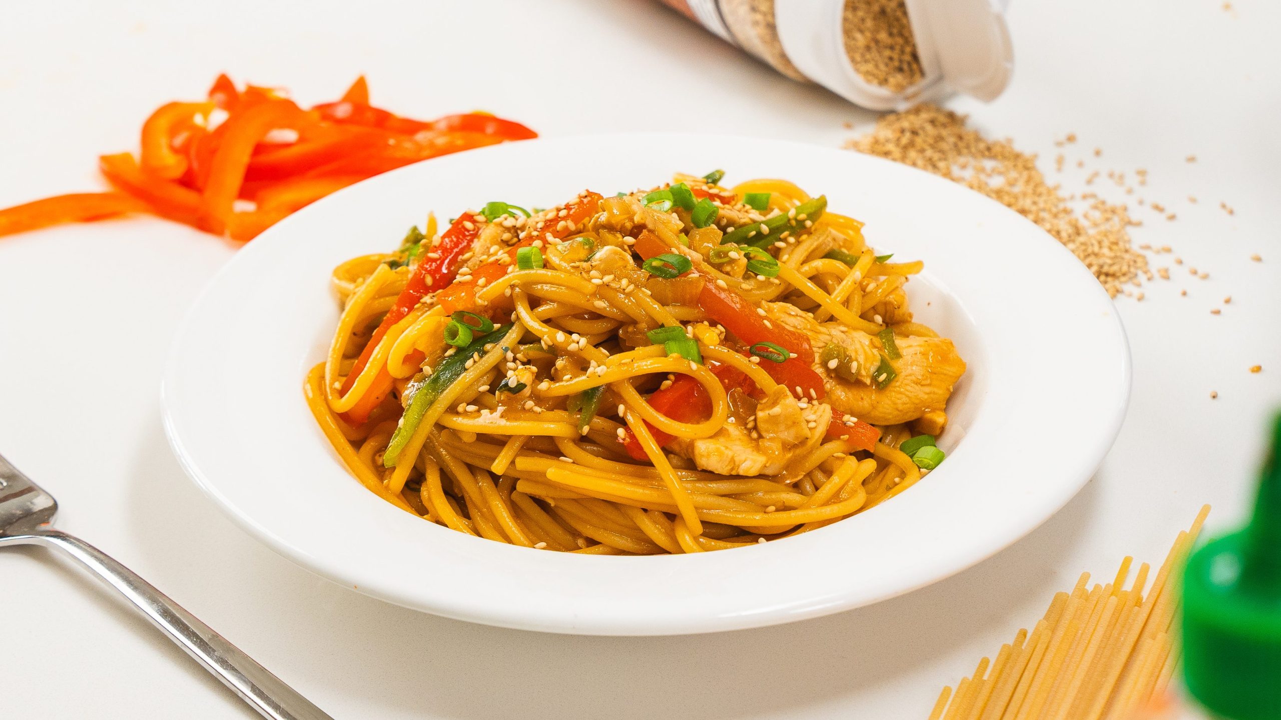 Stir fry in a white bowl. Snap peas, red pepper and dry pasta is visible to the side.