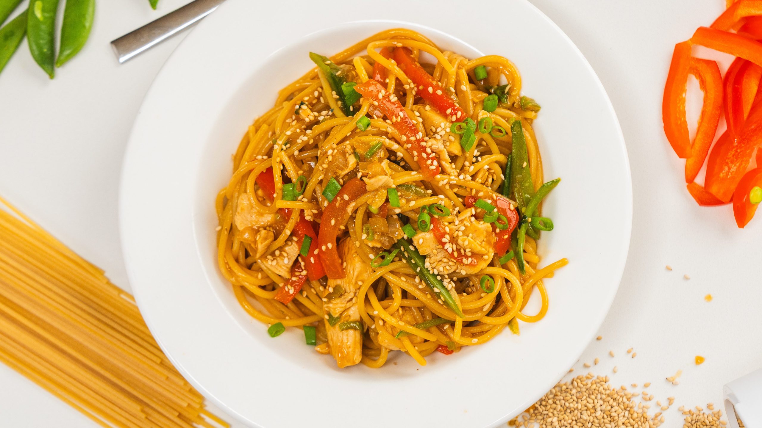 Chicken noodle stir fry in a white bowl, garnished with sesame seeds and green onion.