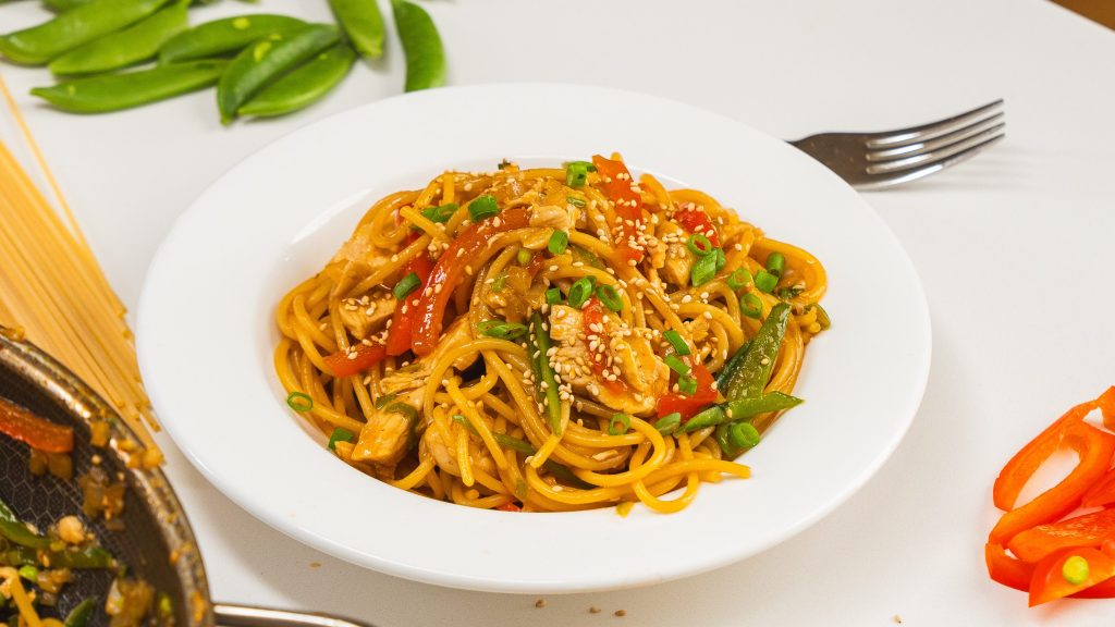 One pan chicken noodle stir fry recipe in a white bowl with green onion and sesame seeds on top.