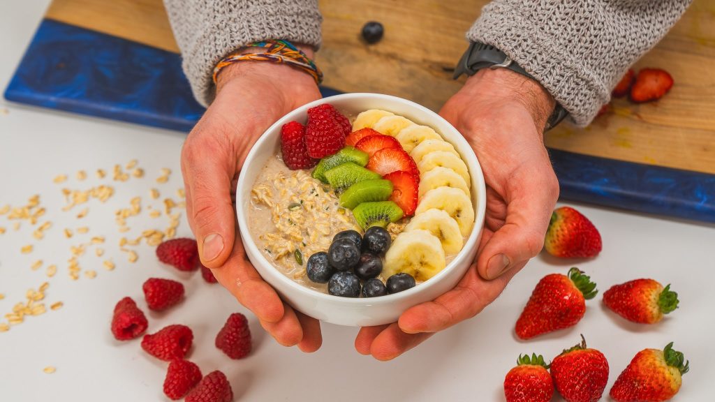 Hands holding a white bowl of protein overnight oats topped with berries, bananas and kiwi. A spoon and fresh fruit are visible in the background.