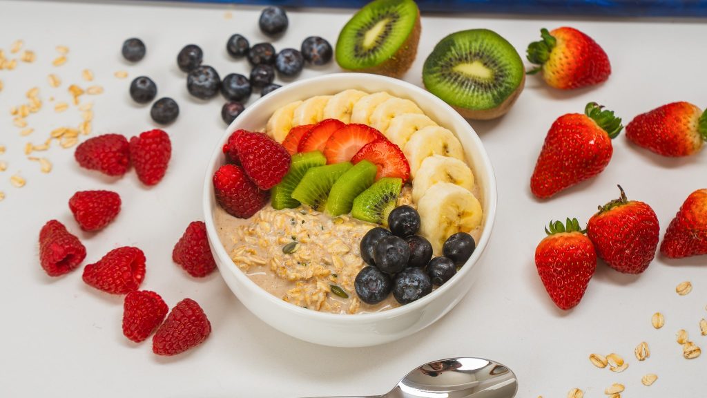 a white bowl of protein overnight oats topped with berries, bananas and kiwi. A spoon and fresh fruit are visible in the background.