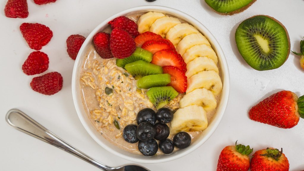 a white bowl of protein overnight oats topped with berries, bananas and kiwi. A spoon and fresh fruit are visible in the background.