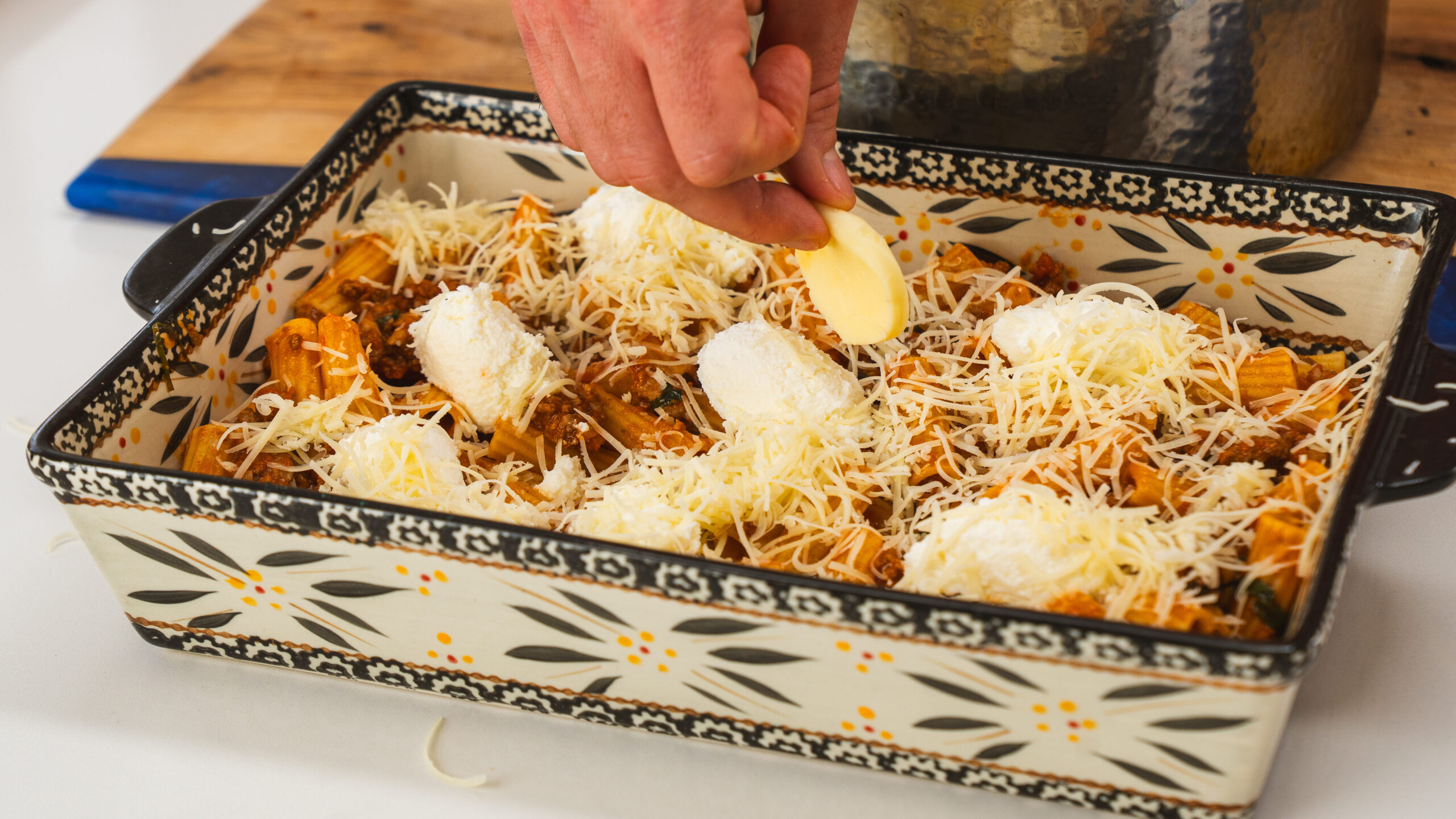 Based rigatoni being assembled in a colourful casserole dish, with fresh mozzarella and parmesan cheeses being sprinkled on top.