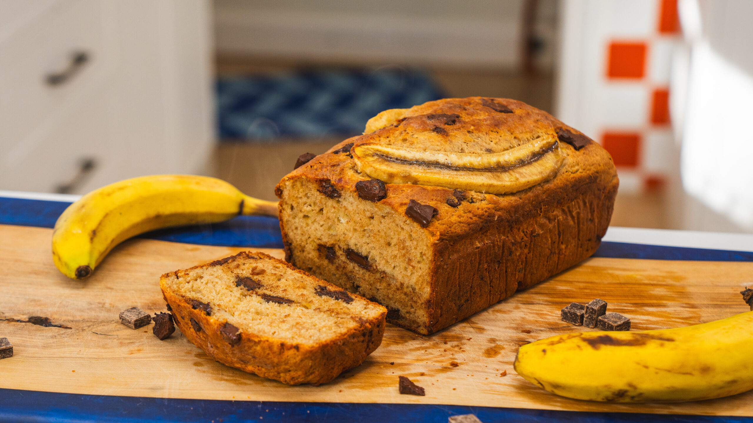 Cottage cheese banana bread sitting on a wooden counter with bananas and chocolate chunks scattered to the side.