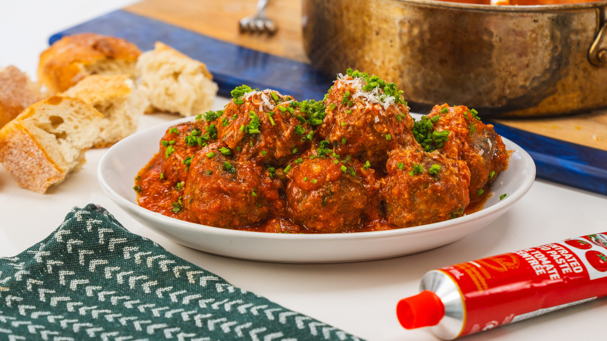 meatballs and tomato cause in a white bowl on a counter top. A tea towel and tube of tomato paste is visible from the side.