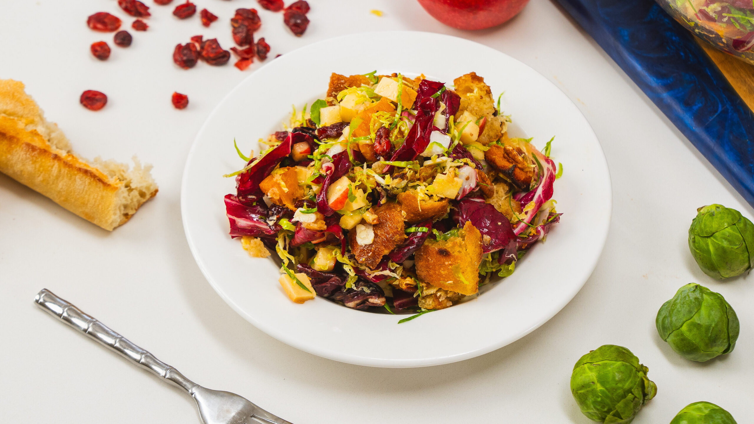 A winter panzanella salad in a bowl. Dried cranberries, bread and Brussels sprouts are visible to the side.