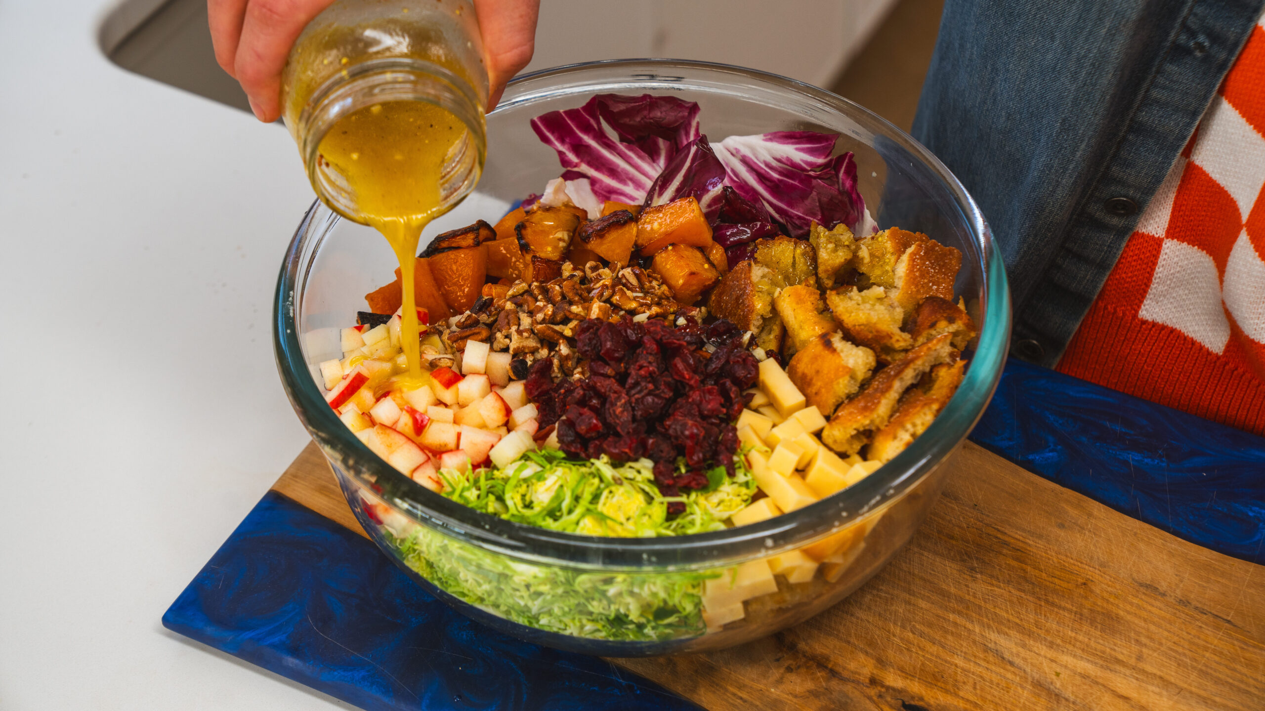 A bowl filled with a winter panzanella salad: Brussels sprouts, dried cranberries, apple, roasted squash with dressing being poured on top.