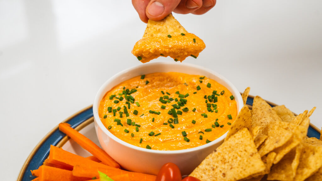 Cottage cheese ranch dip with buffalo sauce in a bowl topped with chives and fresh veggies on the side. Someone is taking a scoop out of the bowl using a nacho chip.