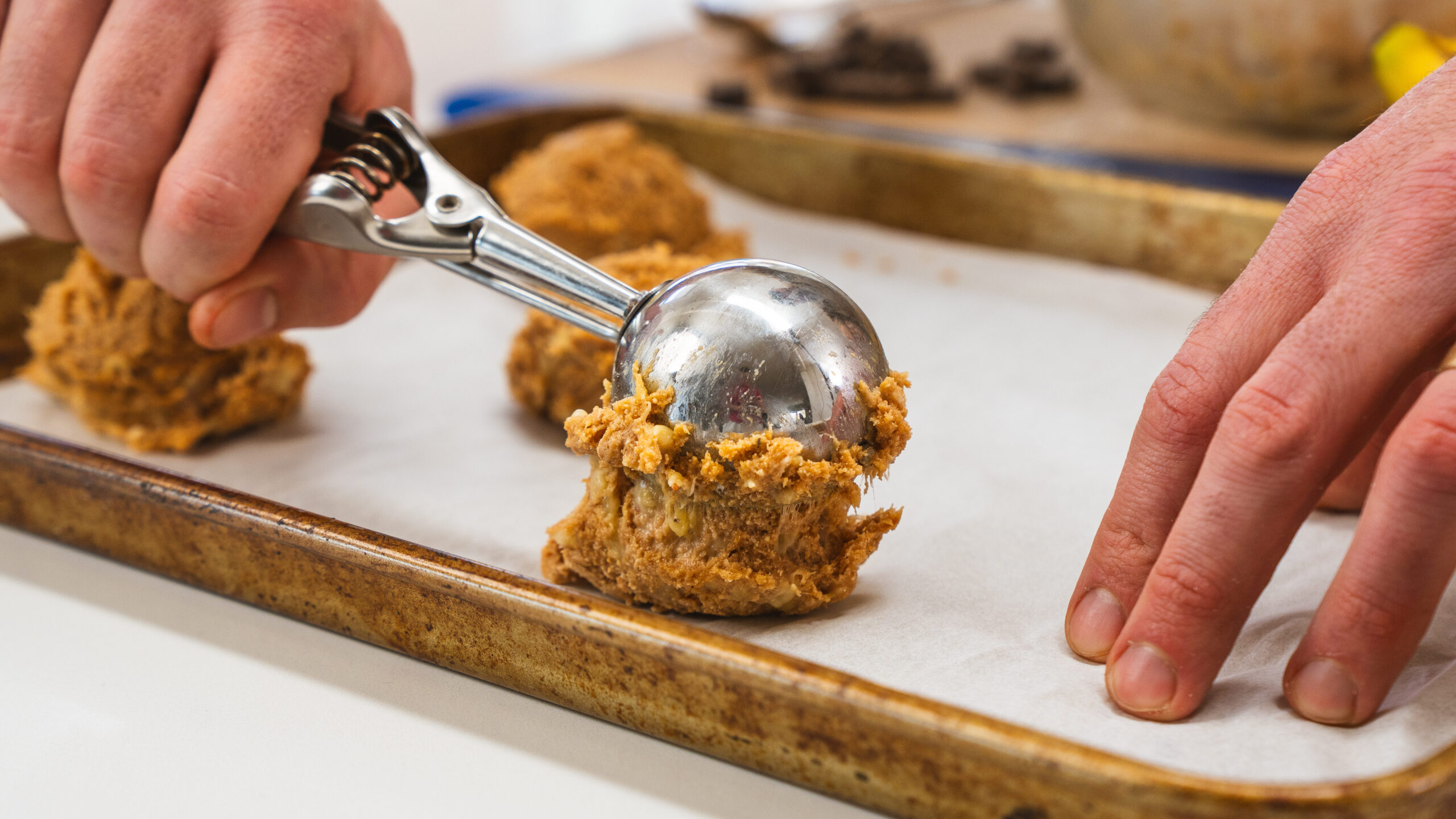Protein chocolate chip cookie batter being placed on a parchment-lined cookie sheet with an ice cream scoop.