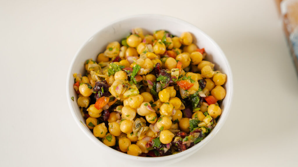 A chickpea salad with colourful veggies and fruit in a white bowl on a counter.
