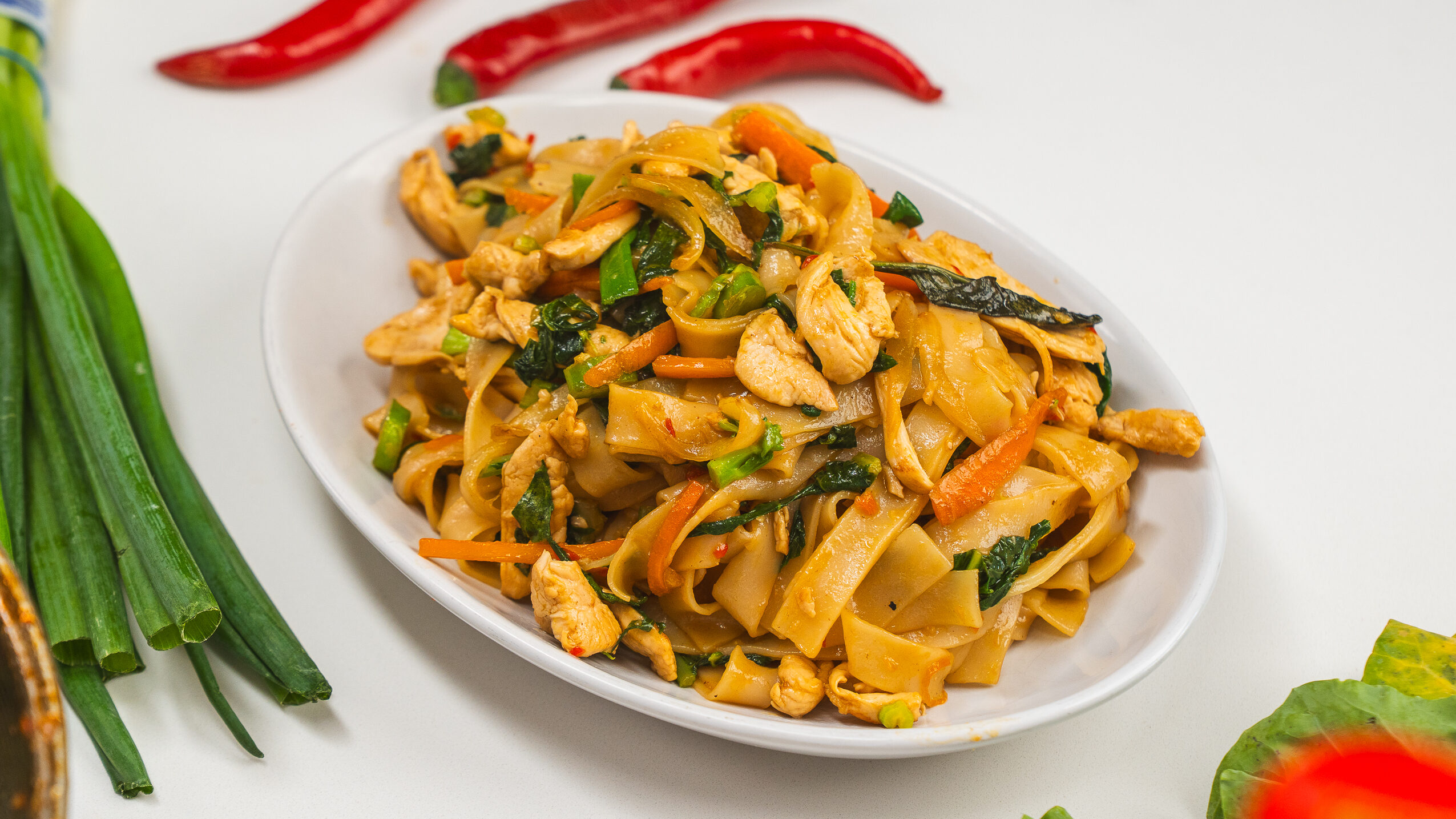 Thai drunken noodles on a white plate with thai red chilis in the background.
